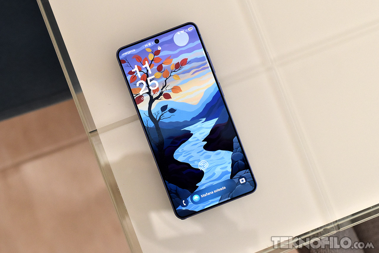 Smartphone sobre mesa con fondo de pantalla de paisaje otoñal y río serpenteante, hora y fecha visibles.