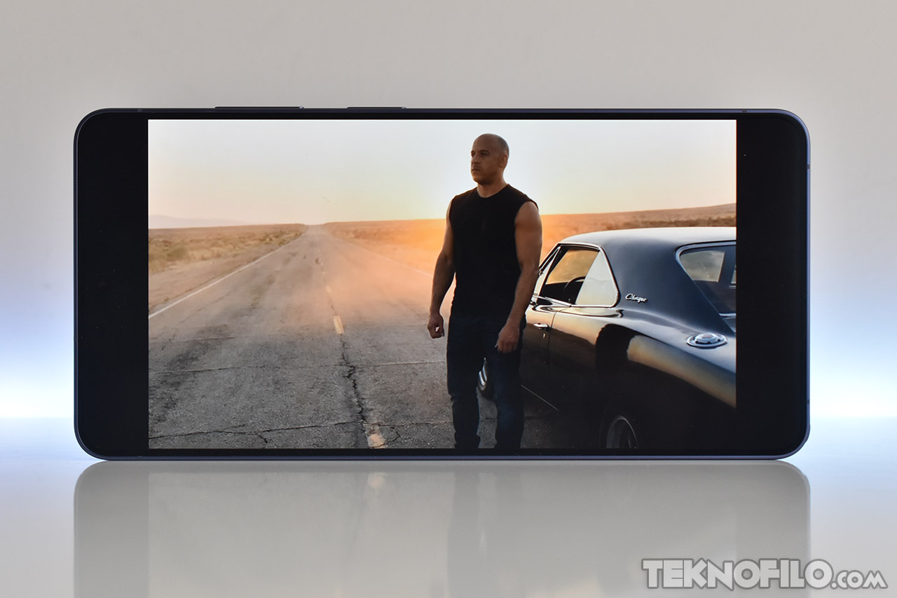 Hombre en camiseta negra de pie junto a un coche clásico en una carretera desierta al atardecer, visto en pantalla.