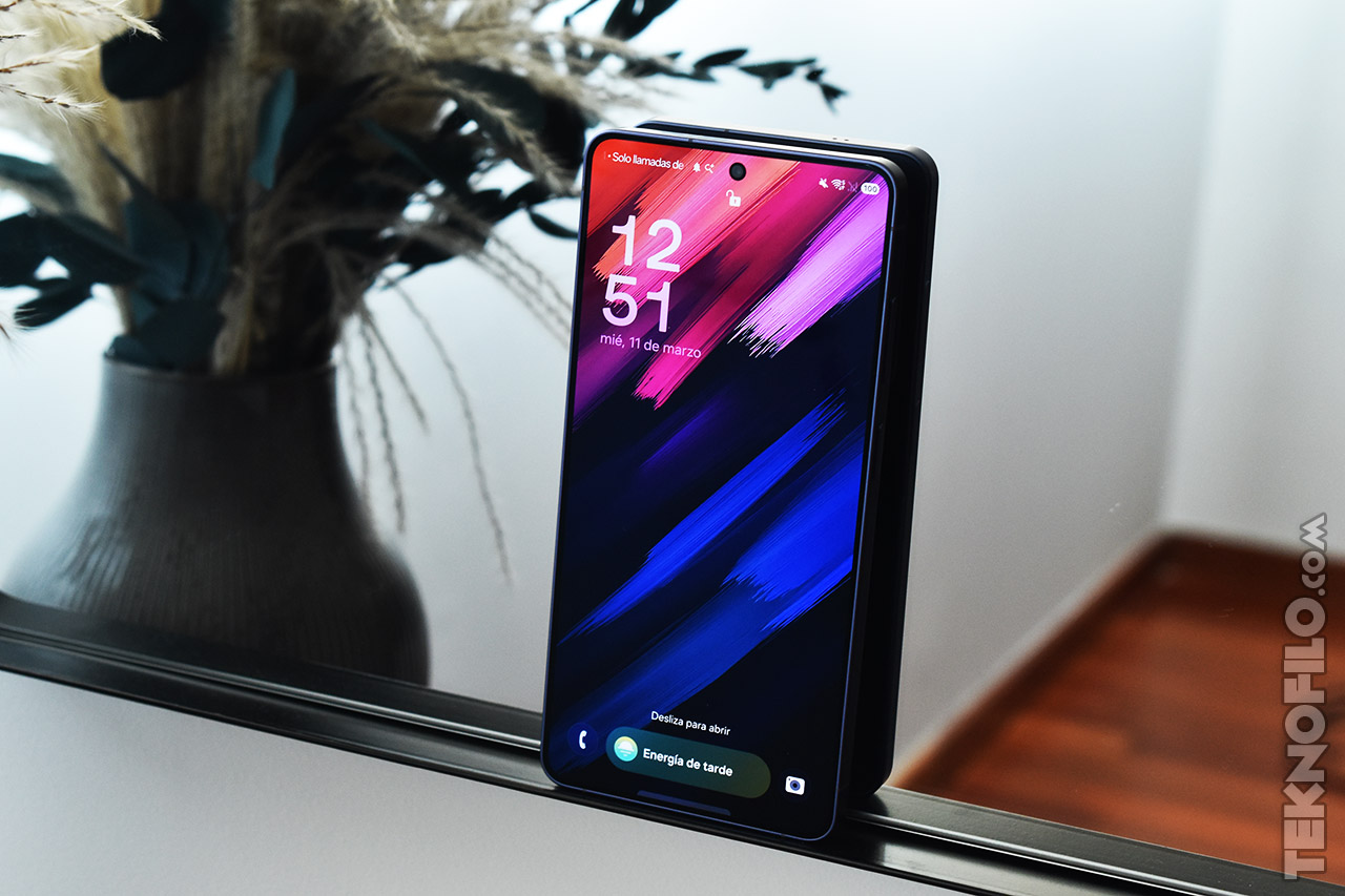 Smartphone con pantalla de inicio colorida sobre base de vidrio, junto a planta decorativa.