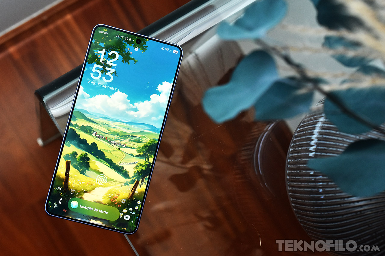 Smartphone con pantalla encendida mostrando paisaje animado, sobre mesa de cristal junto a decoraciones.