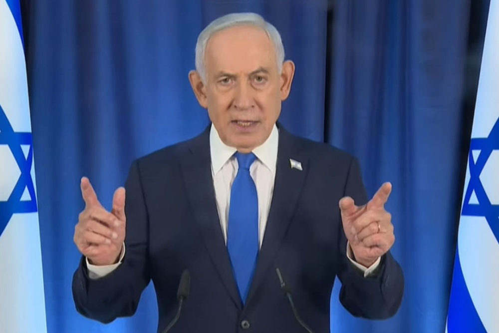 El vídeo de Netanyahu con “seis dedos” desata el caos: ¿Ha sido sustituido por una IA? Hombre en traje dando un discurso frente a banderas israelíes.