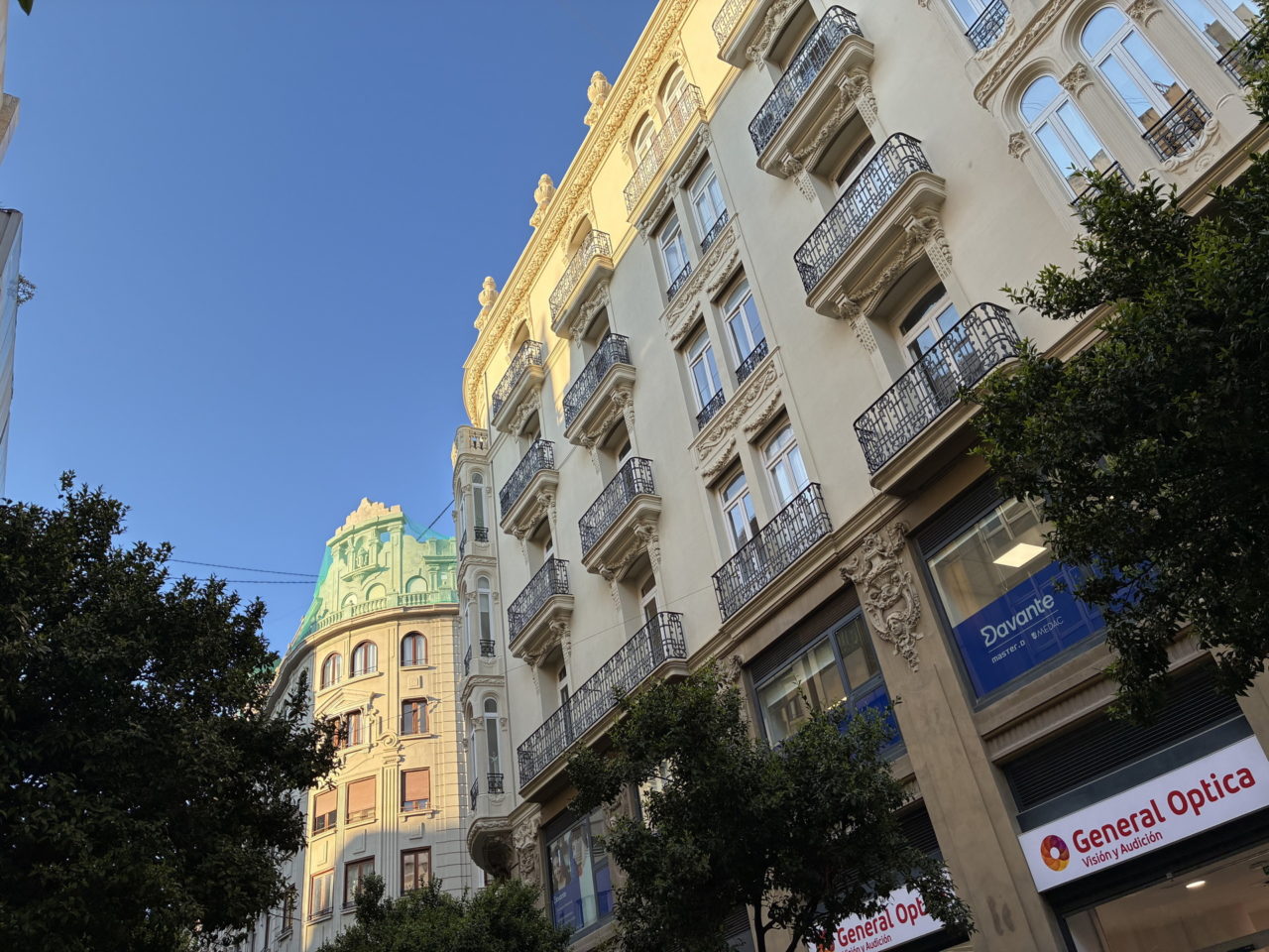 Fachadas de edificios históricos bajo un cielo azul en el centro de una ciudad, con tiendas en el nivel inferior.