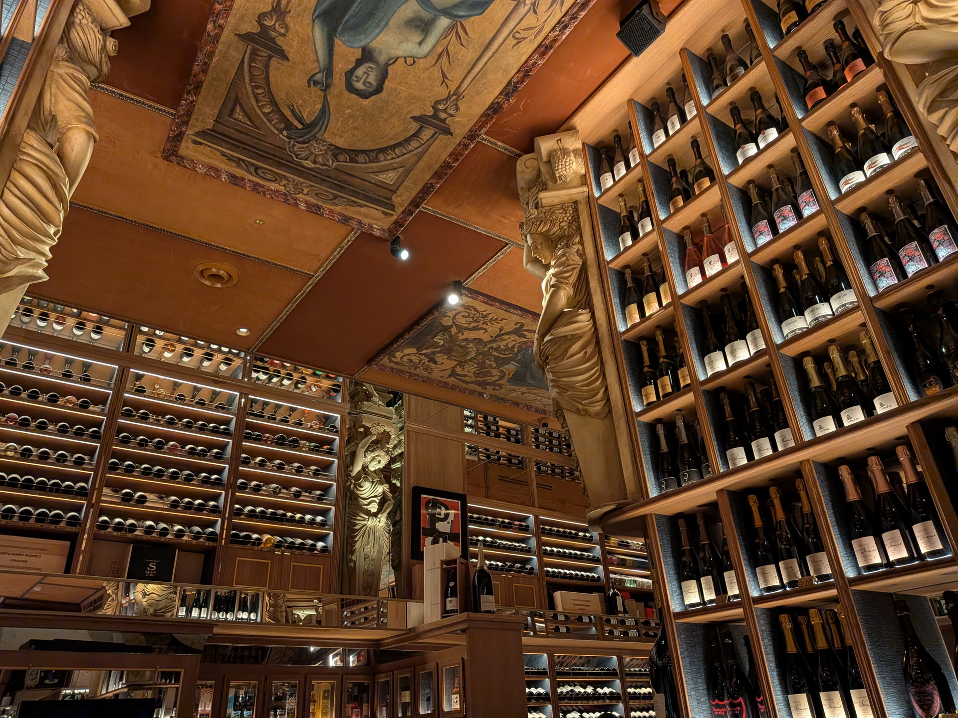 Bodega elegante con estanterías de vinos y techo decorado con arte clásico. Ambiente cálido y sofisticado.