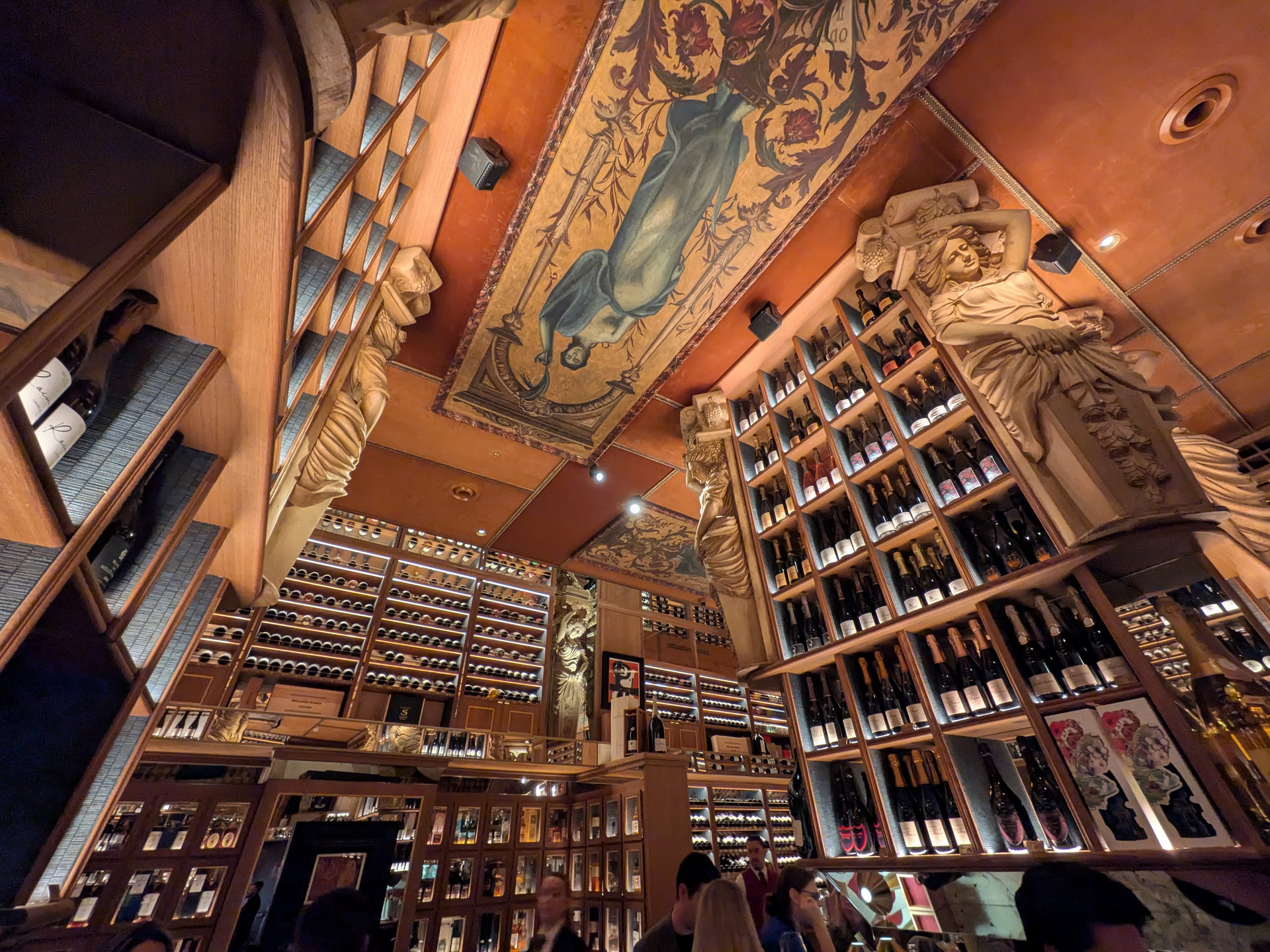 Interior de una bodega con estantes llenos de botellas de vino y decoración clásica en las paredes y techo.
