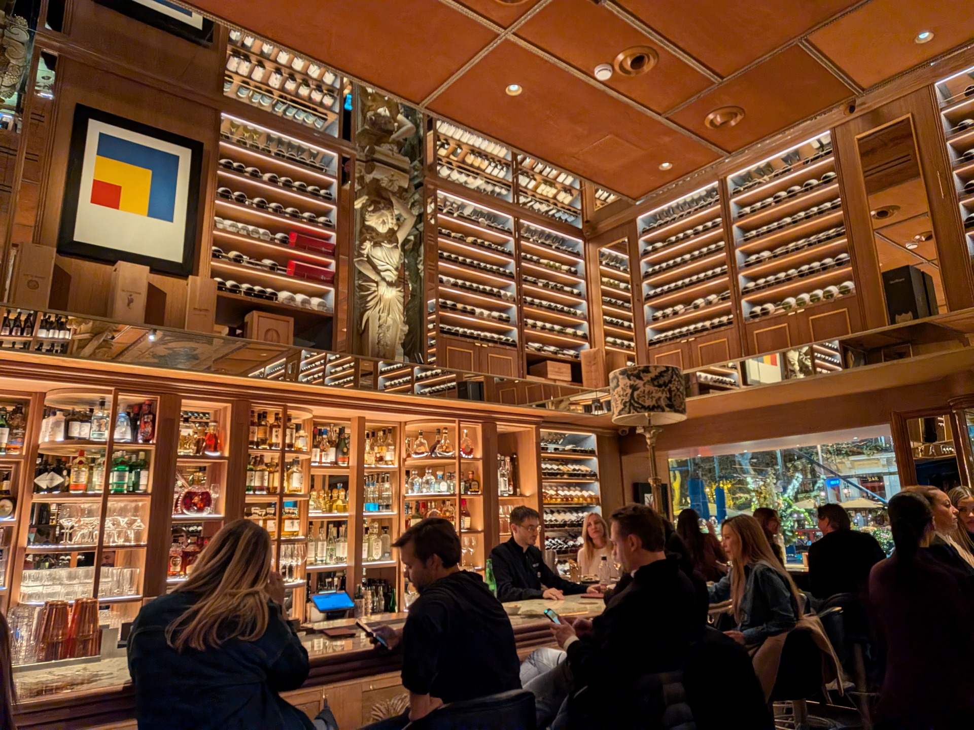 Bar moderno con estantes de vinos iluminados y clientes charlando en mesas, creando un ambiente acogedor.