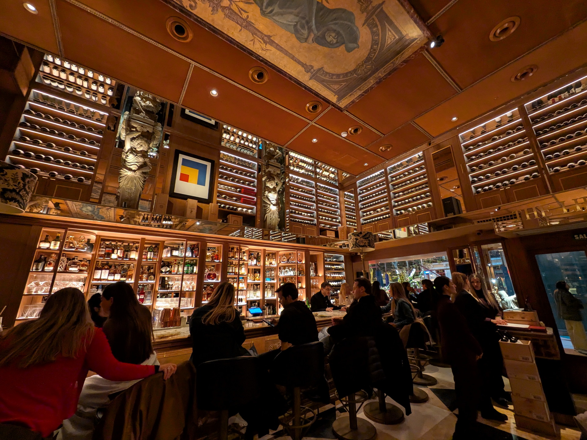 Bar elegante con estantes llenos de botellas y arte en el techo, gente disfrutando de bebidas en un ambiente acogedor.