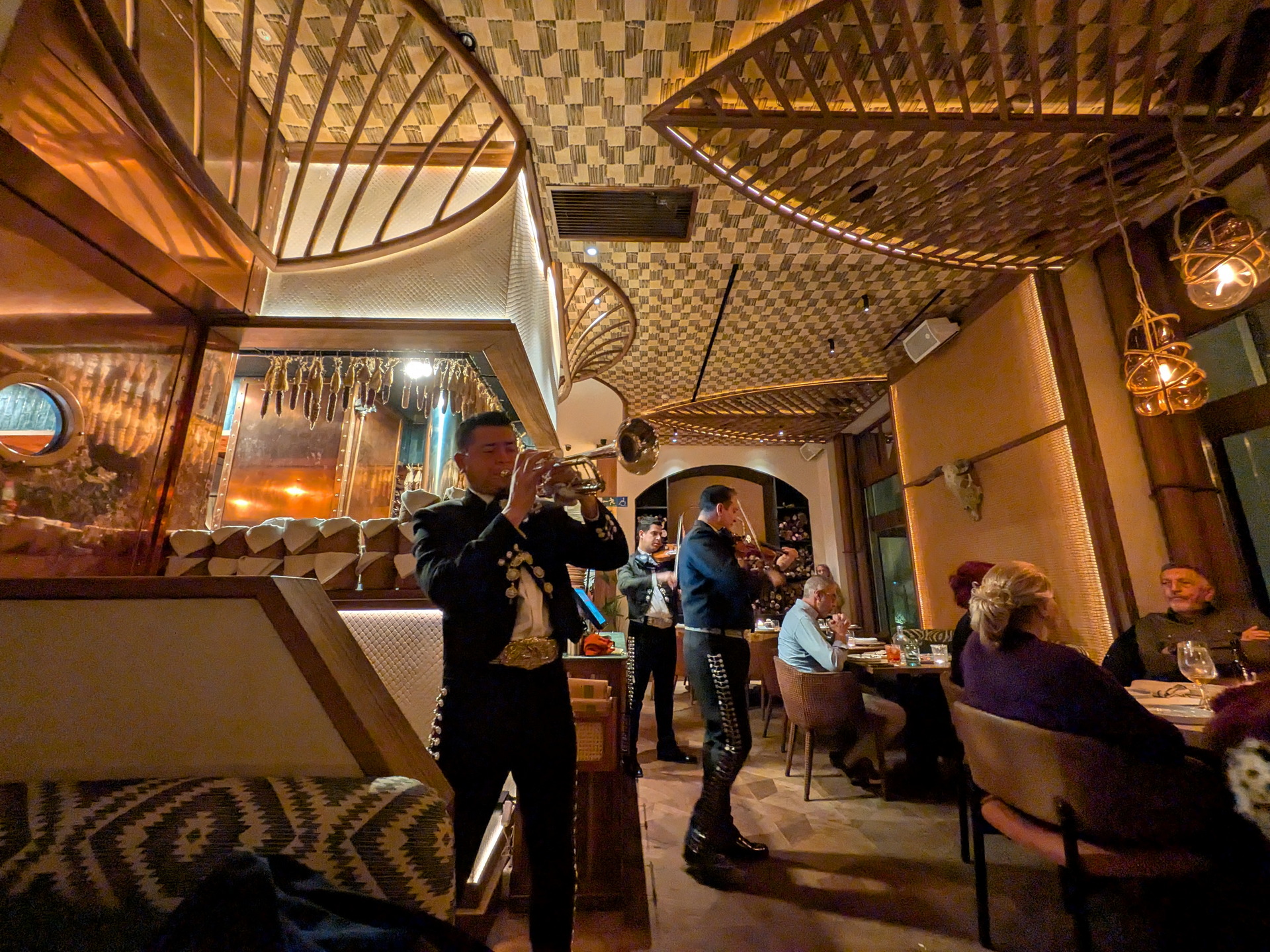 Mariachi tocando trompeta en restaurante elegante con comensales disfrutando ambiente acogedor. Decoración sofisticada.