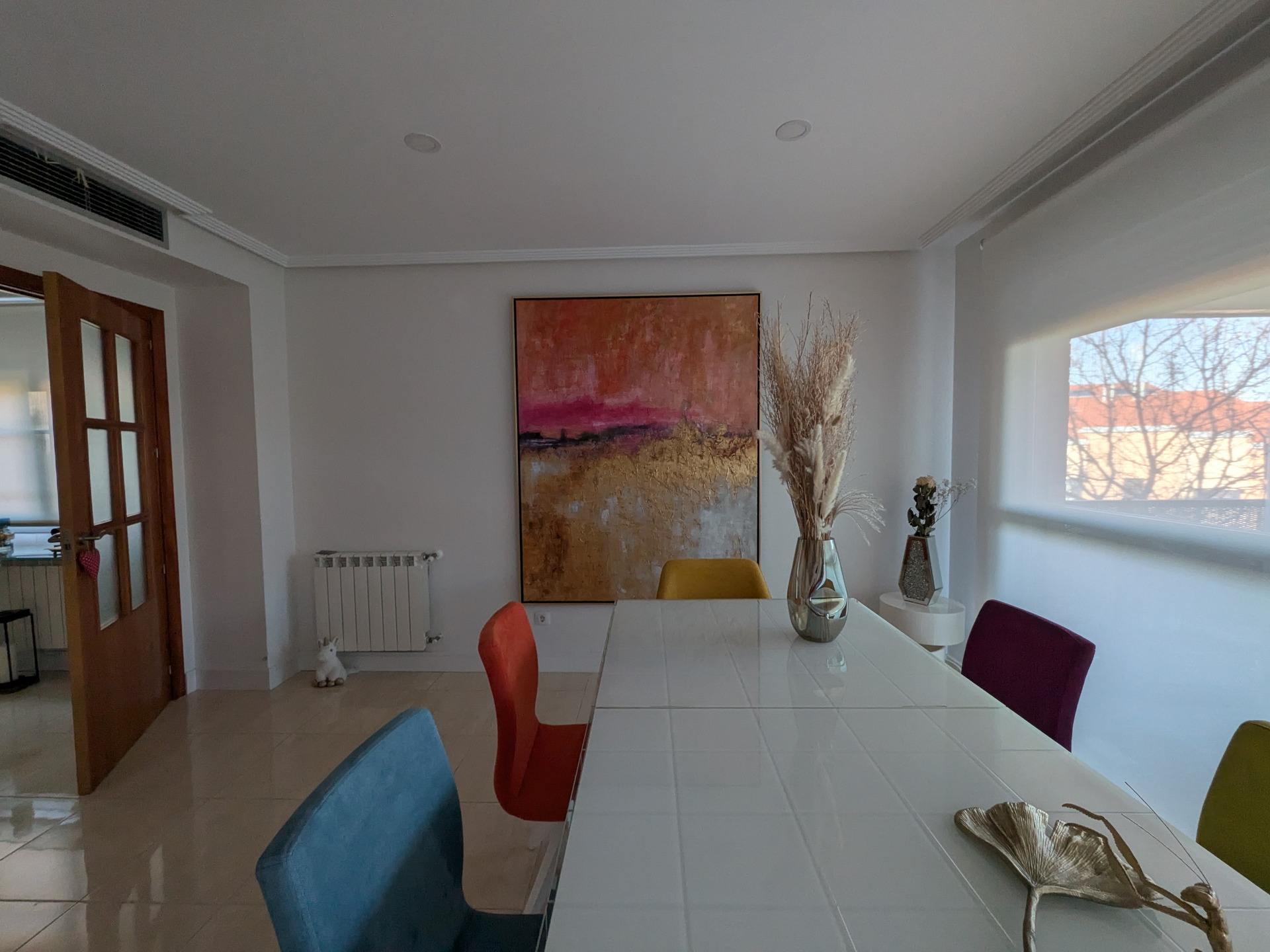 Comedor moderno con sillas coloridas, mesa blanca, cuadro abstracto y jarrón decorativo junto a ventana luminosa.