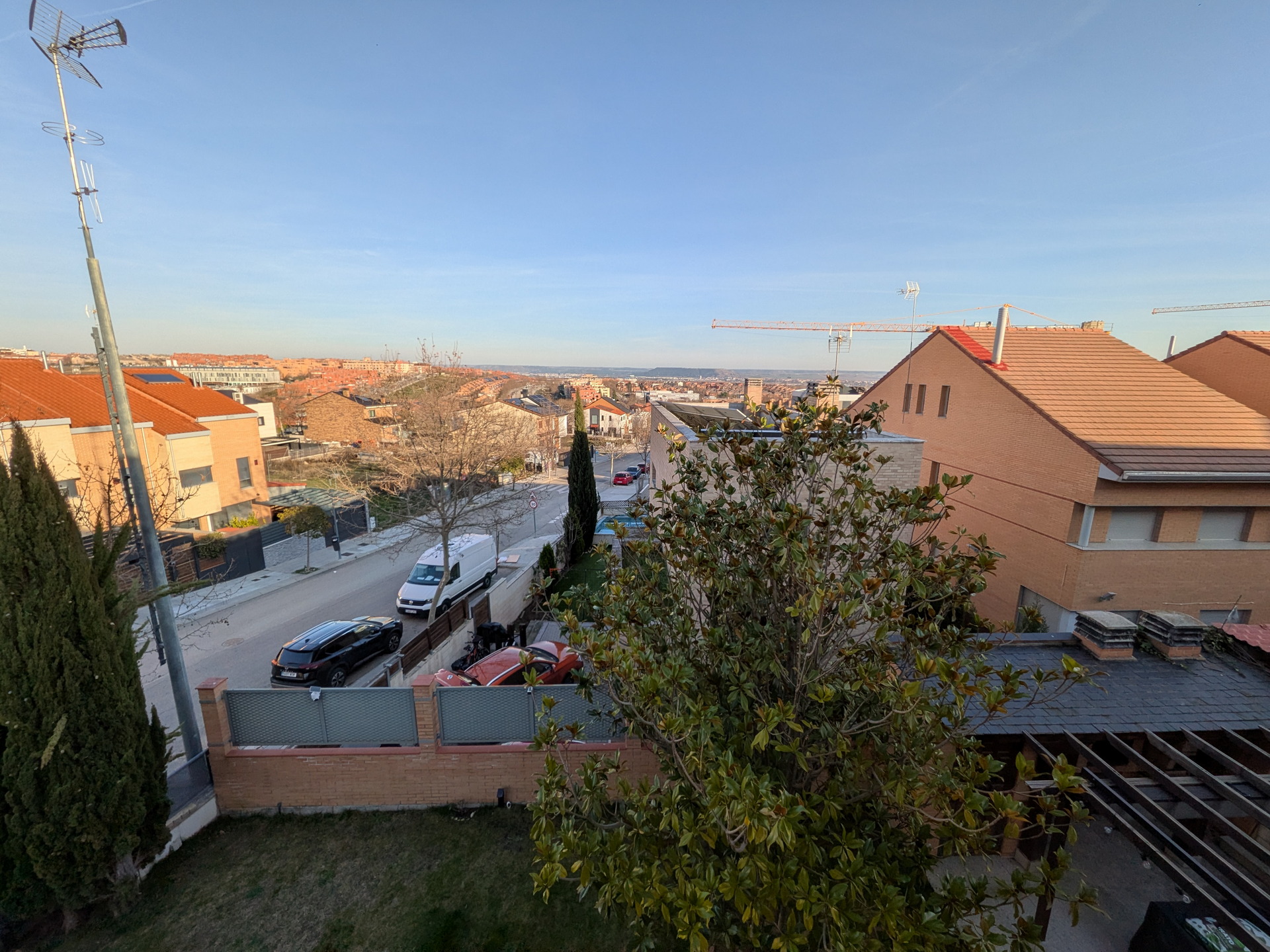 Vista urbana de un barrio residencial con casas, antenas y grúas bajo un cielo despejado al atardecer.