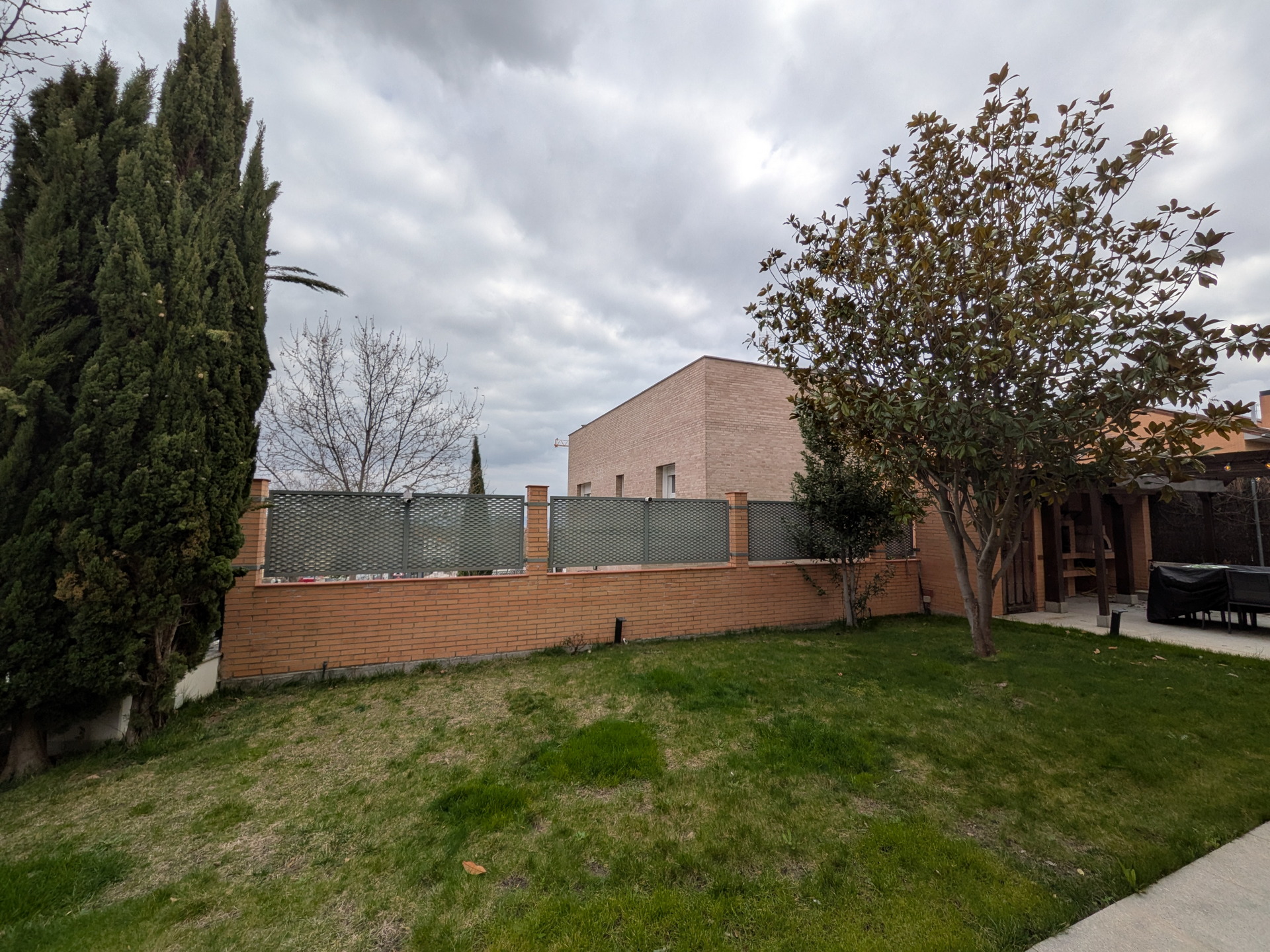 Jardín con césped y árboles frente a una casa de ladrillo y cielo nublado.