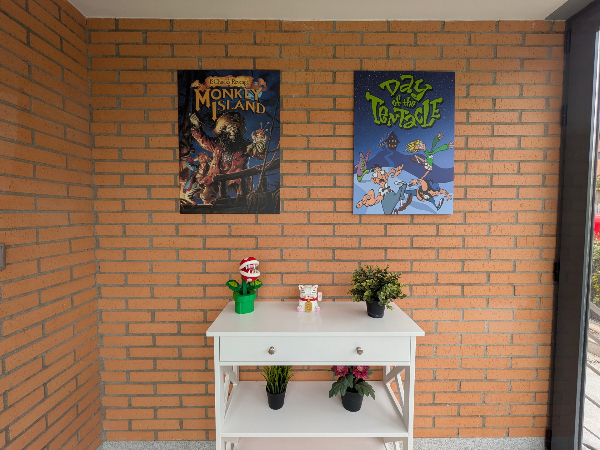 Pared de ladrillo con pósters de videojuegos retro sobre mesa blanca decorada con plantas y figuras de colección.