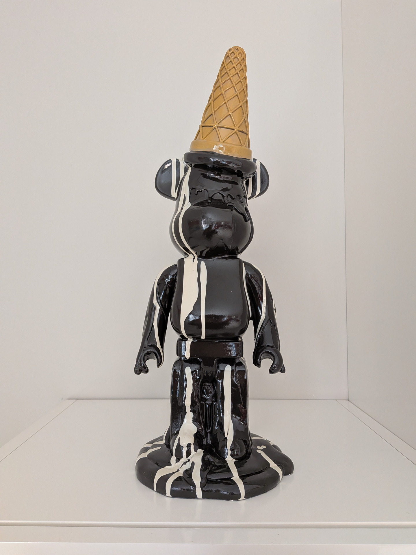 Estatua de oso negro con sombrero de cono de helado y manchas blancas, sobre estante blanco minimalista.