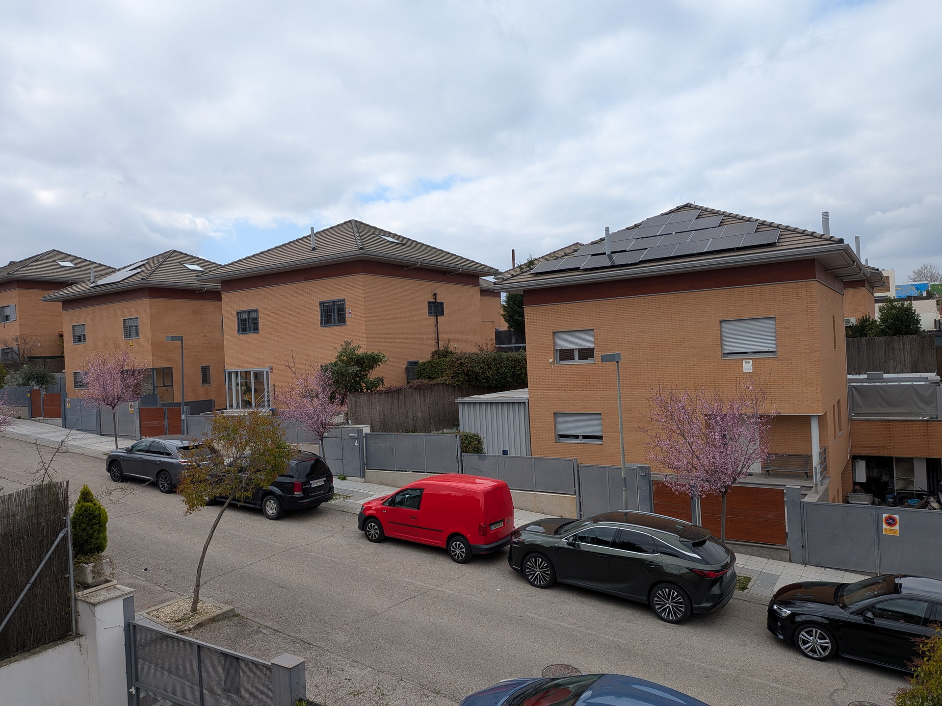 Casas de ladrillo con paneles solares en el techo y árboles en flor, coches estacionados en la calle.