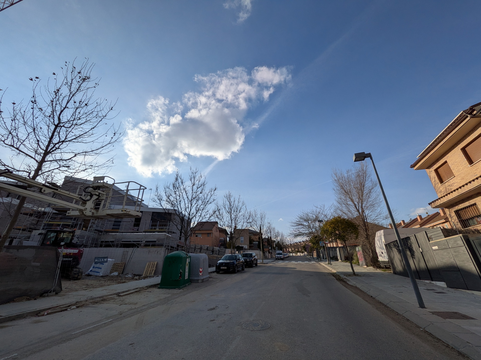 Calle residencial con árboles sin hojas y construcción en proceso bajo un cielo azul con nubes blancas.