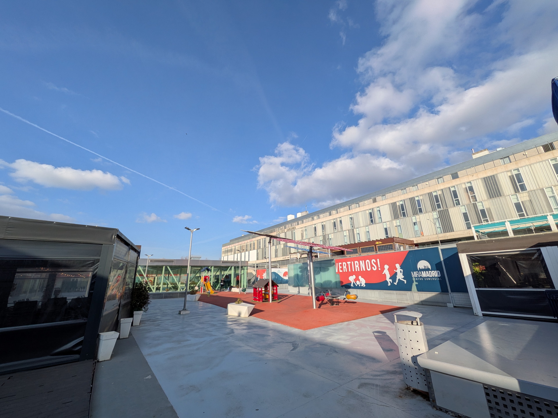 Patio exterior con juegos infantiles, cielo despejado y edificio moderno al fondo en un día soleado.