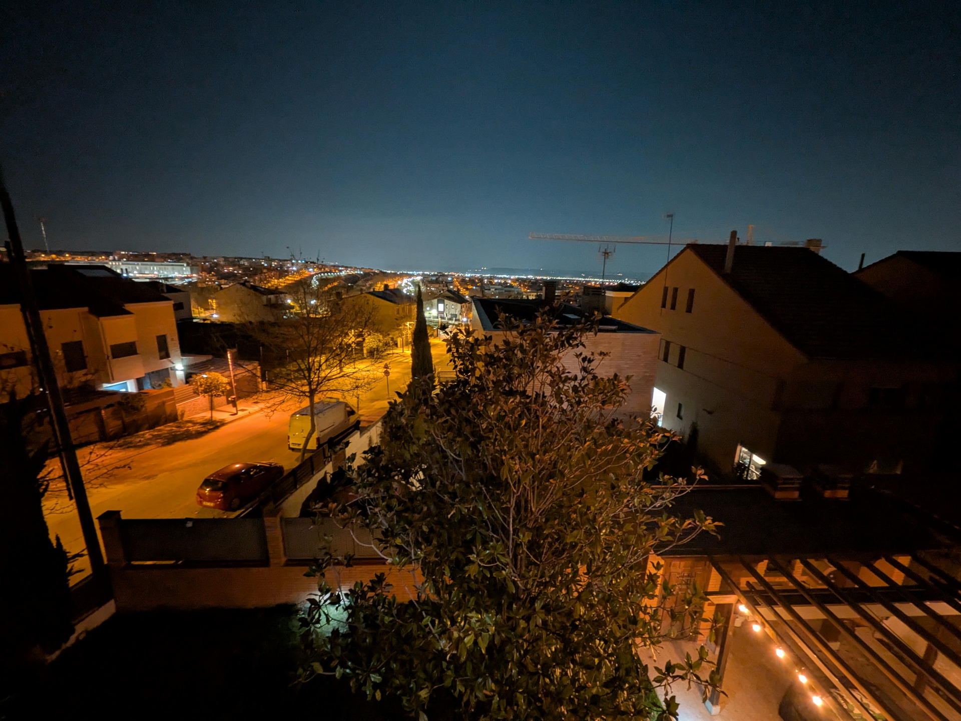 Vista nocturna de una ciudad iluminada desde una terraza, con árboles y luces cálidas en primer plano.