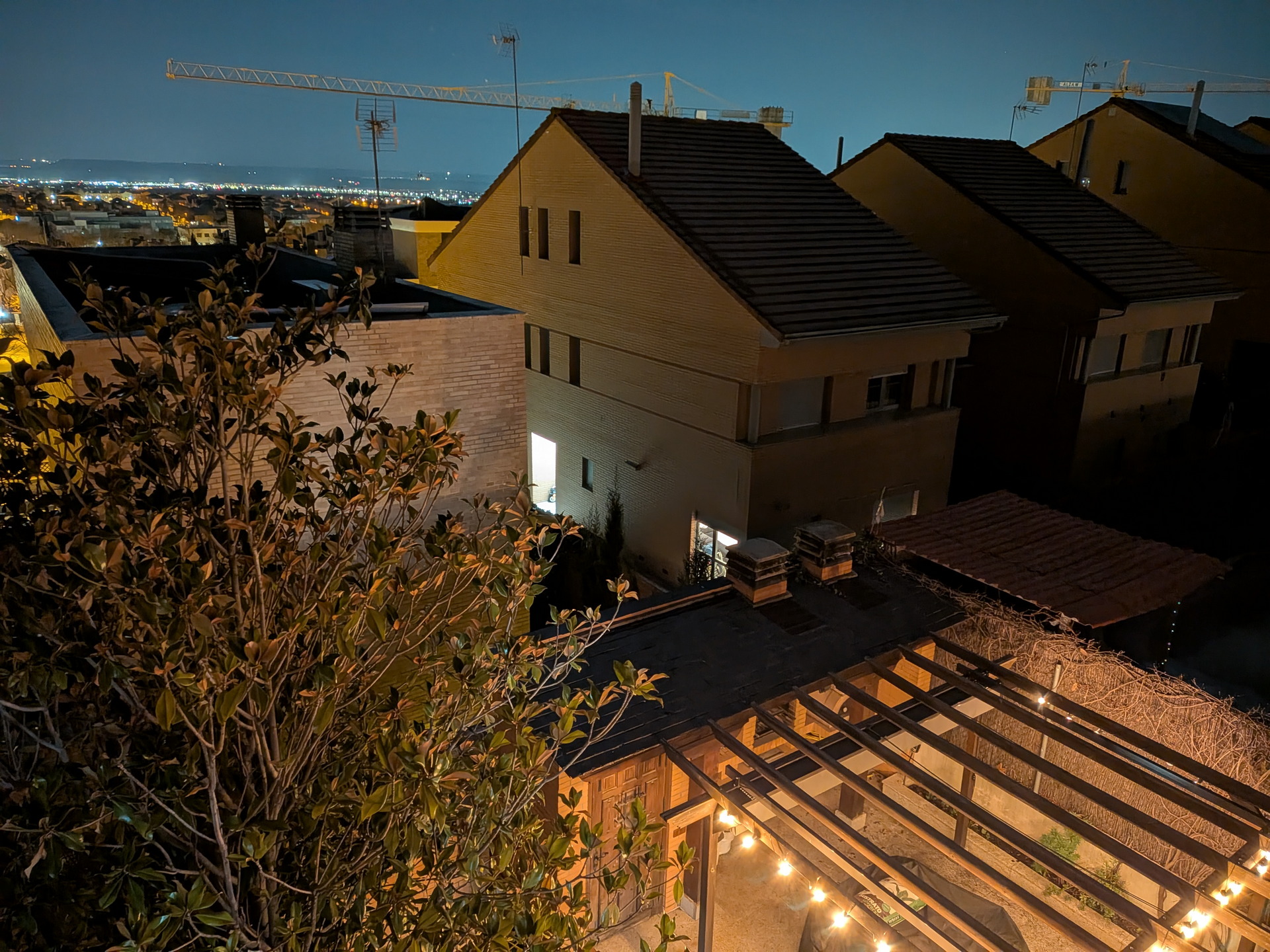 Vista nocturna de casas iluminadas, con grúas al fondo y luces en pérgola en primer plano, cielo despejado.