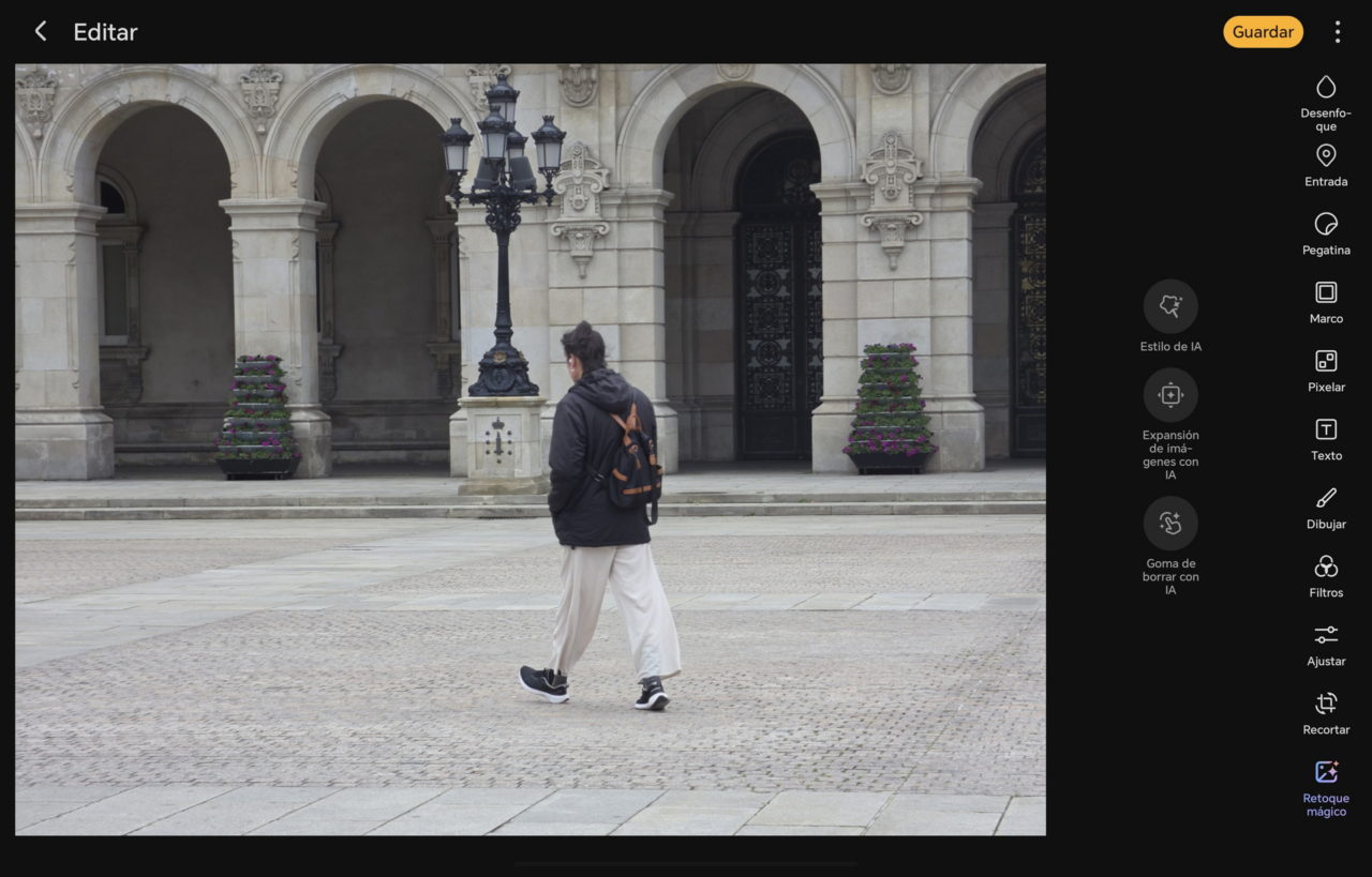 Persona camina frente a edificio histórico con arcos y farola. Lleva chaqueta negra y mochila naranja.