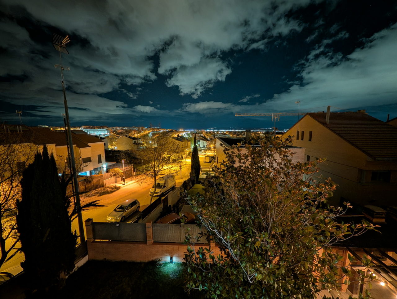 Vista nocturna de una calle urbana iluminada, rodeada de casas y vegetación bajo un cielo nublado.
