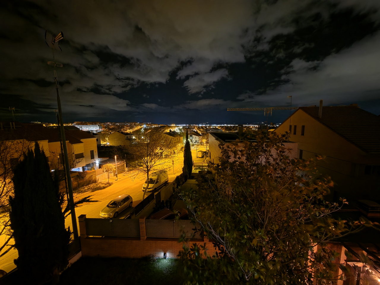 Vista nocturna de un vecindario con calles iluminadas y cielo nublado, resaltando las luces de la ciudad.