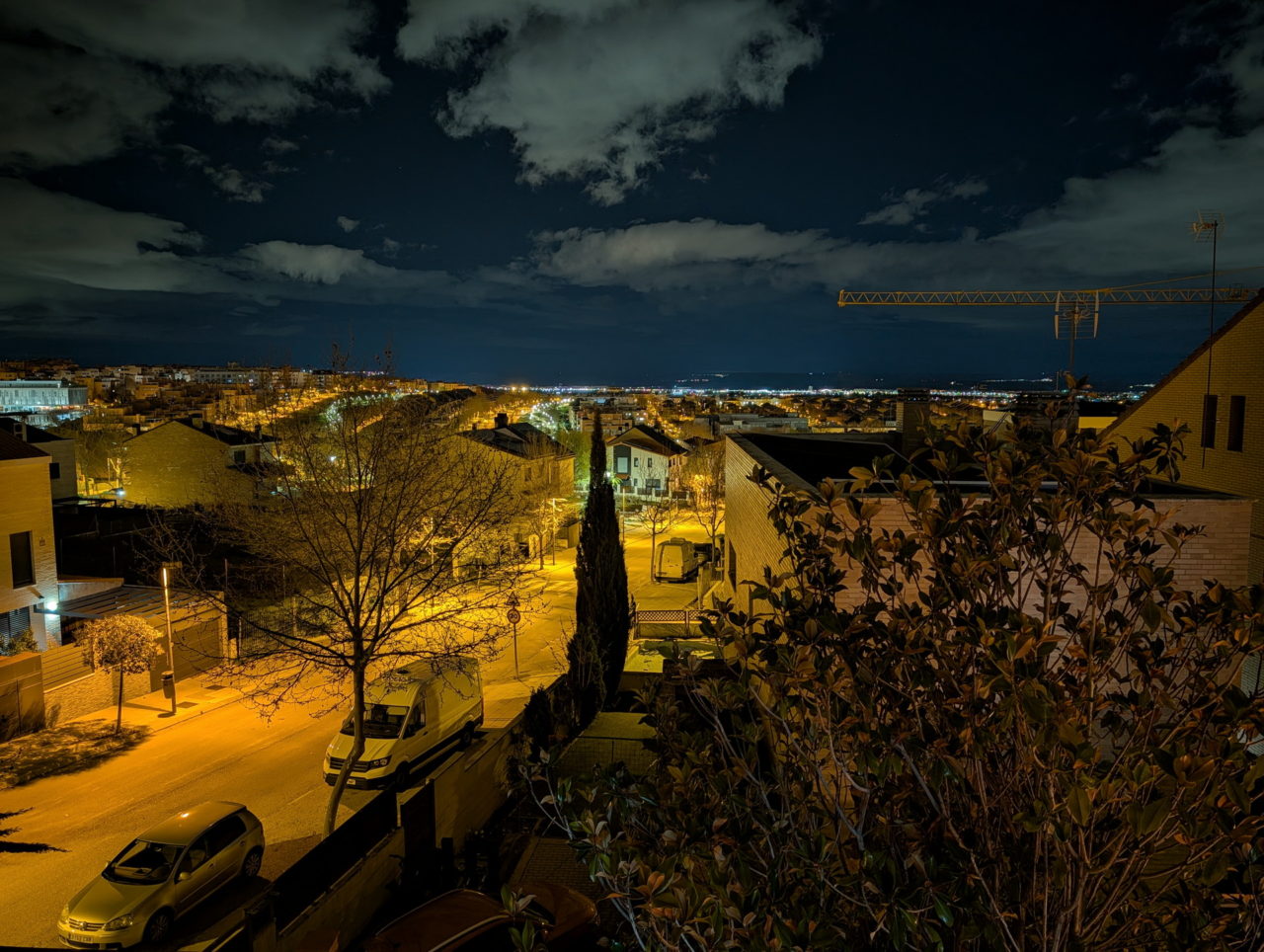 Vista nocturna de un barrio iluminado, con calles y coches visibles, bajo un cielo nublado.