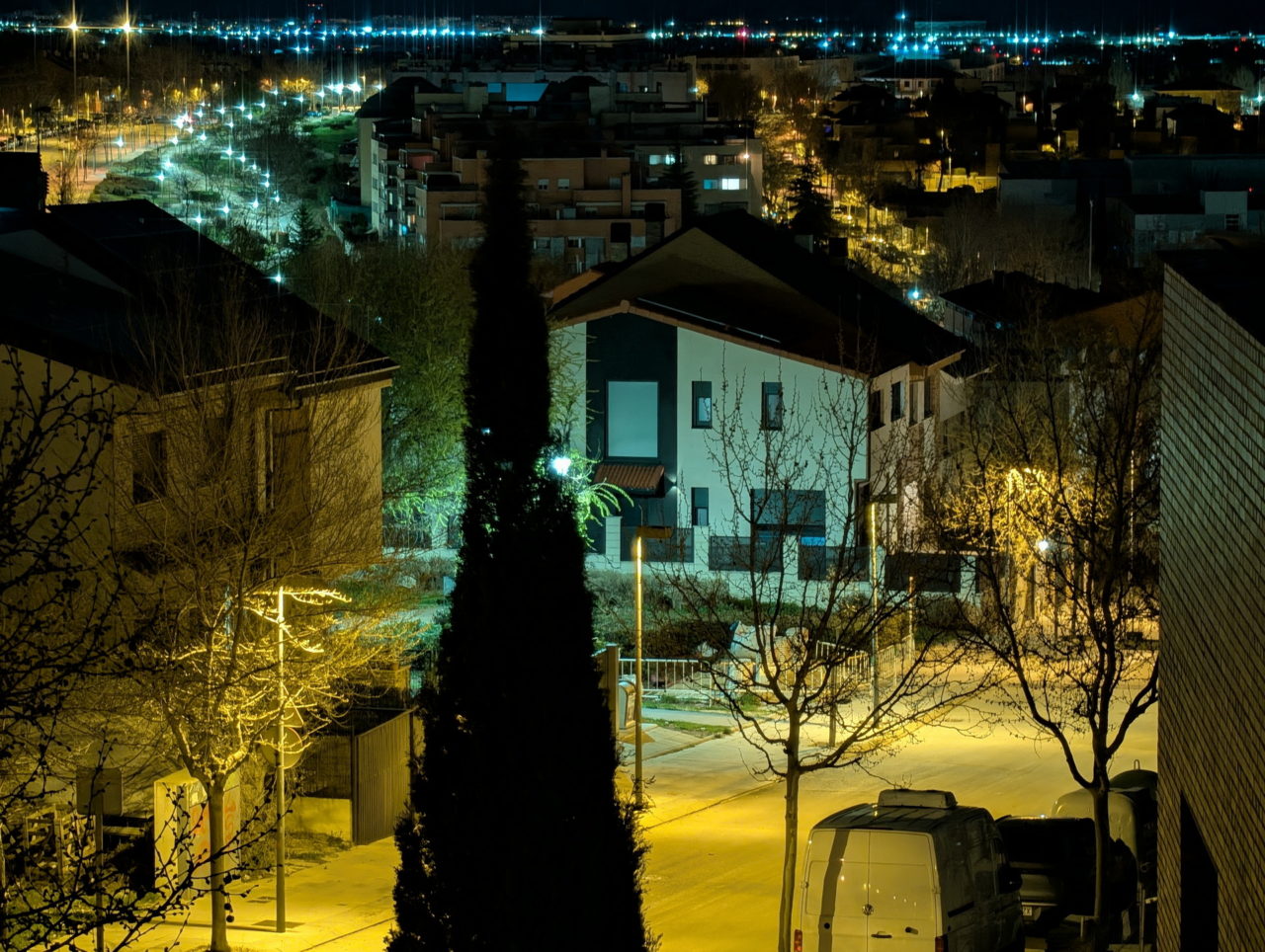 Vista nocturna de un barrio iluminado, con calles vacías y luces brillantes, árboles y casas alrededor.