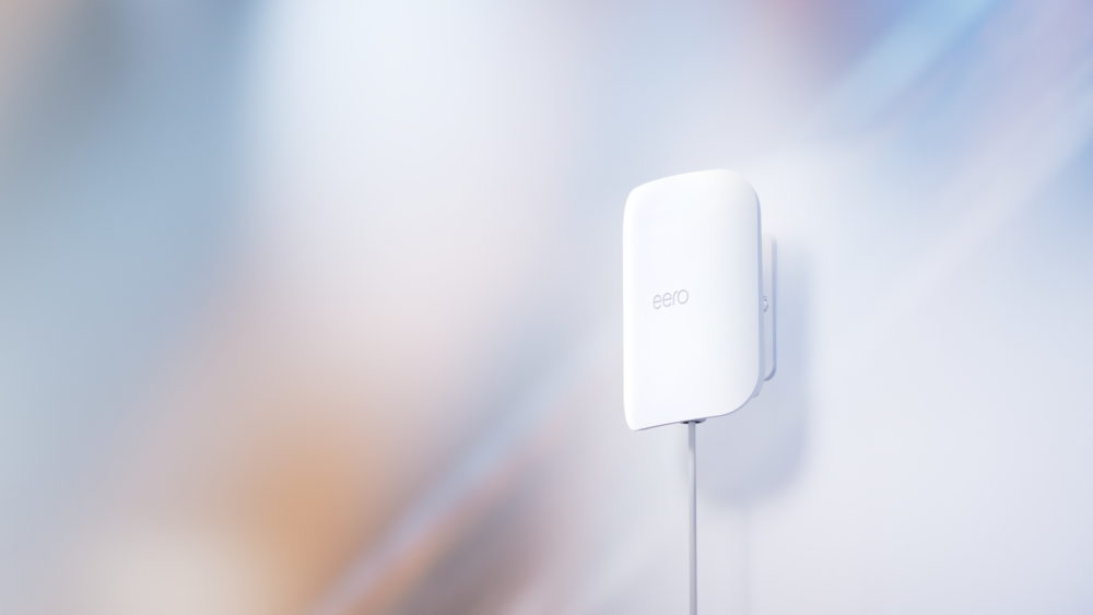 eero Outdoor 7: el nuevo dispositivo que lleva el Wi-Fi 7 al exterior de tu casa Enrutador WiFi Eero blanco montado en la pared, fondo abstracto azul y naranja.