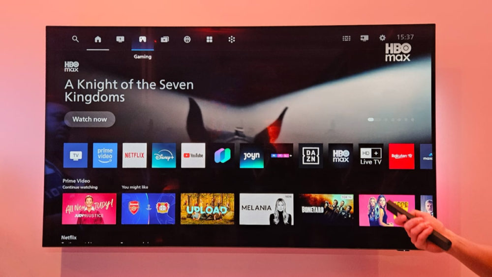 Philips quiere abandonar Google TV para sus televisores inteligentes Pantalla de TV mostrando servicios de streaming como HBO Max, Prime Video, y Netflix, con mando remoto en mano.