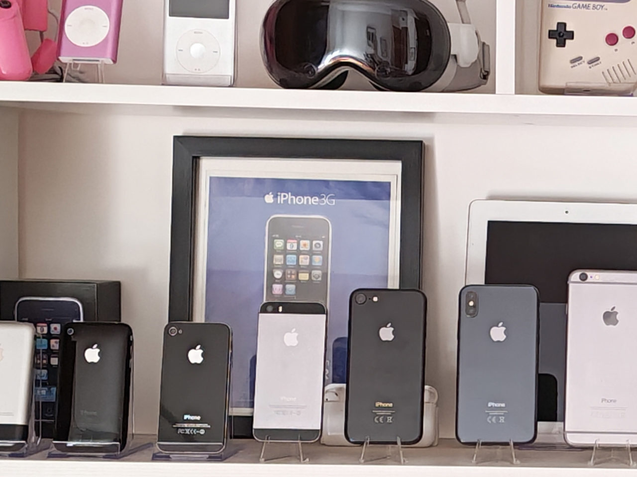 Estantería con colección de iPhones y productos Apple, incluyendo iPods y Game Boy vintage en exhibición.