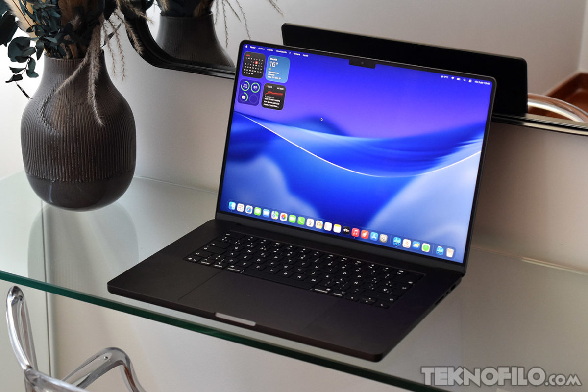 MacBook Pro (2026) review en español: Análisis completo, opinión y prueba real Portátil en una mesa de vidrio con pantalla encendida y fondo azul, al lado de un jarrón decorativo.