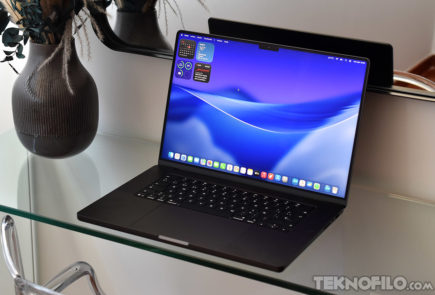 MacBook Pro OLED táctil: Todo apunta a que lo veremos el 28 de octubre Portátil en una mesa de vidrio con pantalla encendida y fondo azul, al lado de un jarrón decorativo.