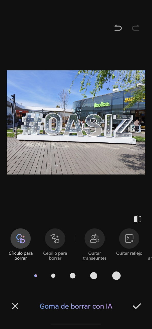 Letrero gigante #OASIZ en centro comercial al aire libre, con tiendas y cielo azul de fondo.