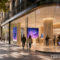 Personas caminando frente a una moderna tienda tecnológica en una calle urbana con árboles y edificios históricos.
