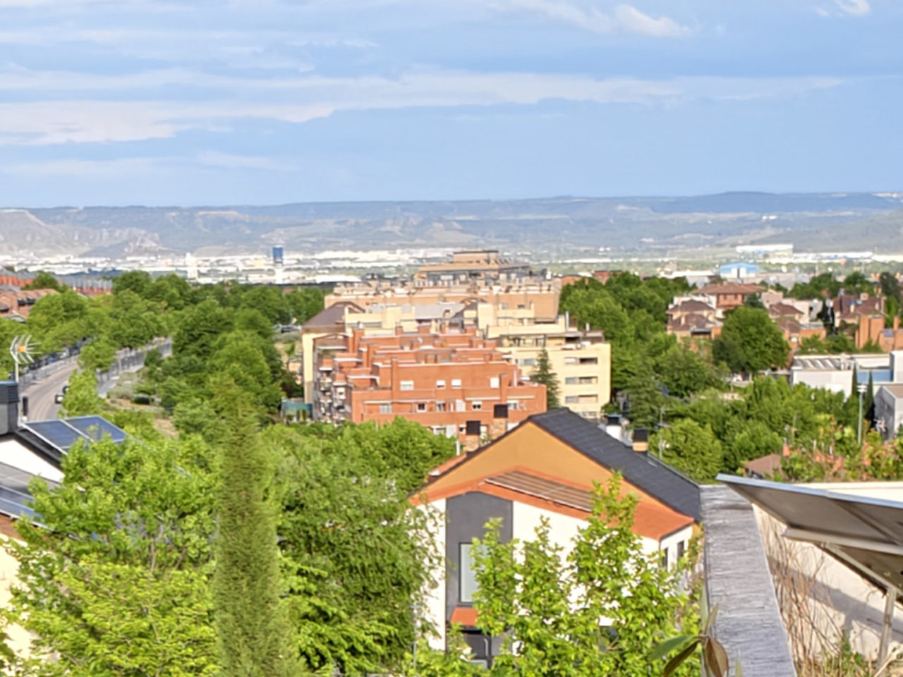 Vista aérea de una ciudad con edificios rodeados de árboles y montañas en el horizonte bajo un cielo despejado.