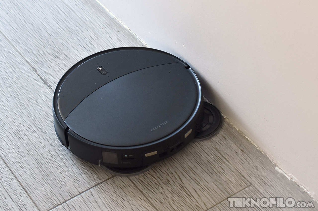 Robot aspiradora negro limpiando junto a la pared en un suelo de madera clara.