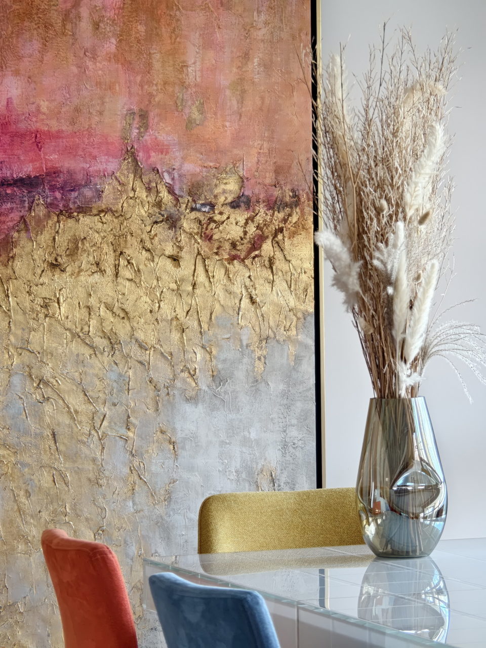 Pintura abstracta dorada y rosa con jarrón de cristal y flores secas sobre mesa moderna, decoración elegante.