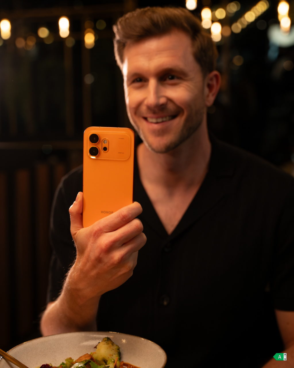 Hombre sonriendo con un teléfono naranja, tomando una foto en un restaurante elegante durante la noche.