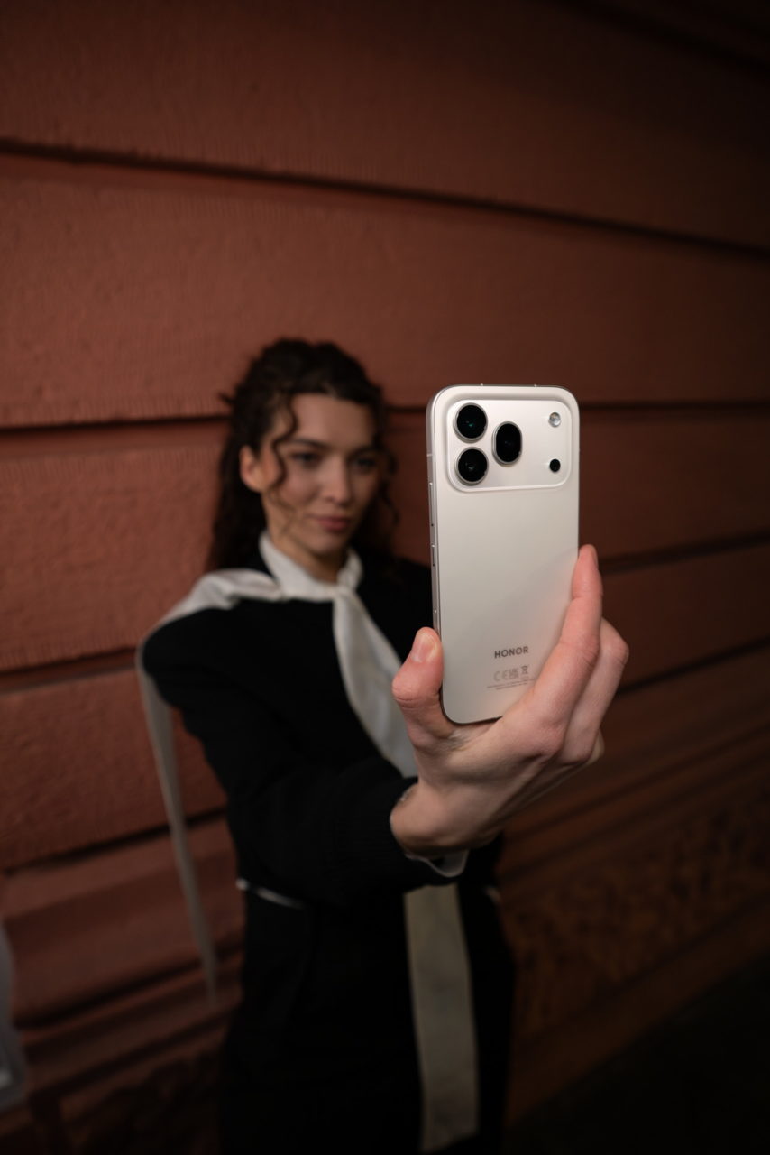 Persona sacándose una selfie con un smartphone HONOR blanco frente a una pared roja.