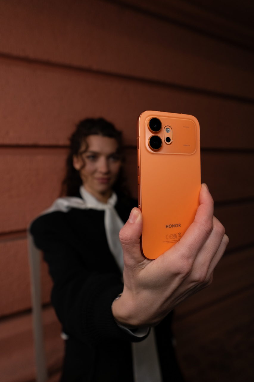 Persona sujetando un teléfono Honor naranja para tomar una selfie, con pared de fondo.