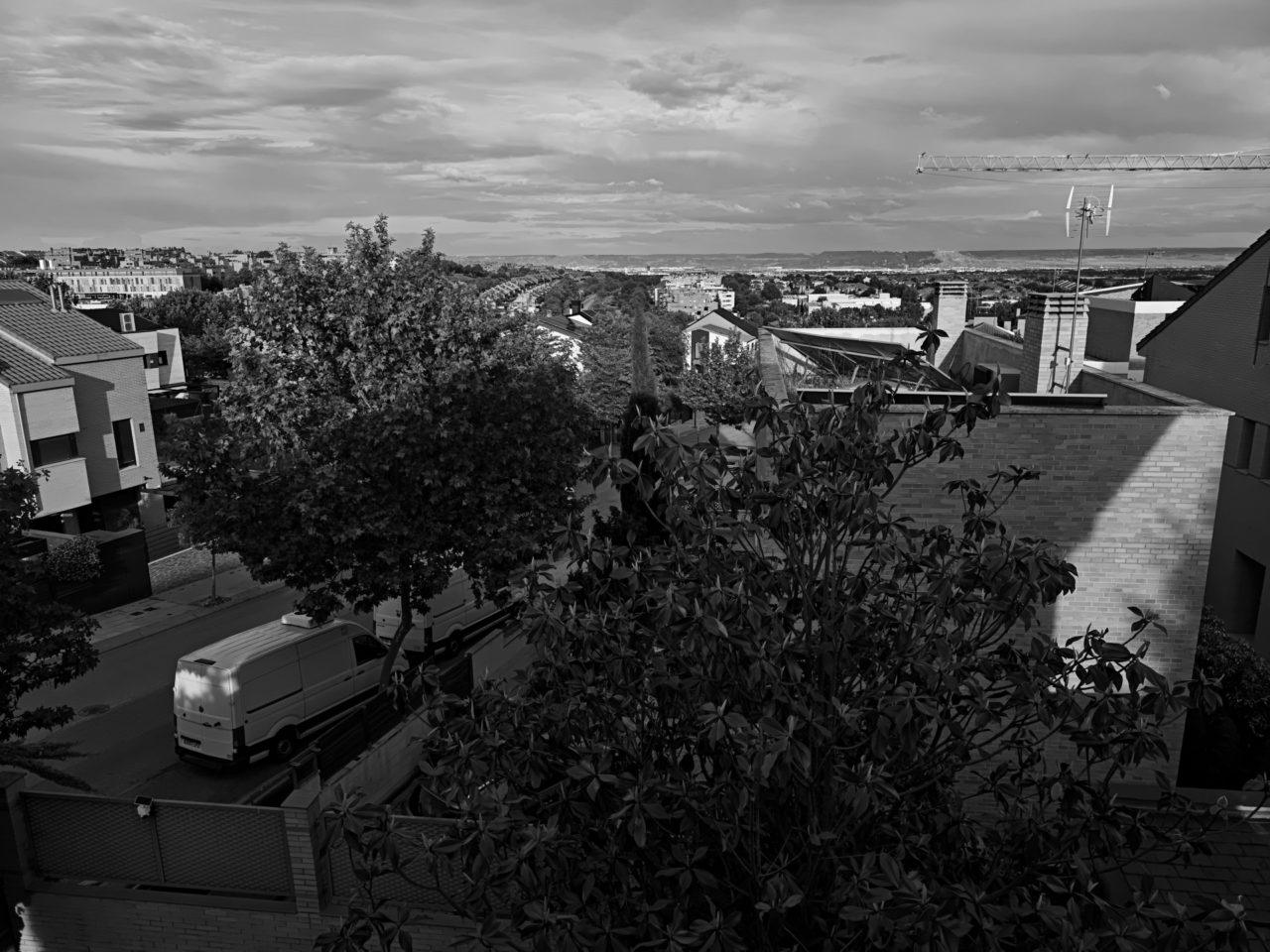 Vista urbana en blanco y negro con casas, árboles y una grúa al fondo en un barrio residencial.