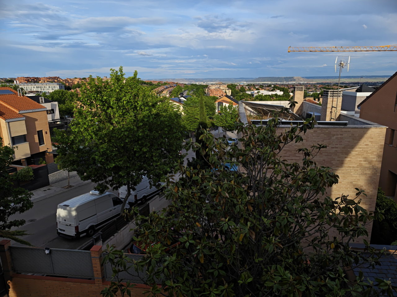 Vista de un barrio residencial con casas de ladrillo, árboles verdes y una grúa en el horizonte bajo un cielo azul.