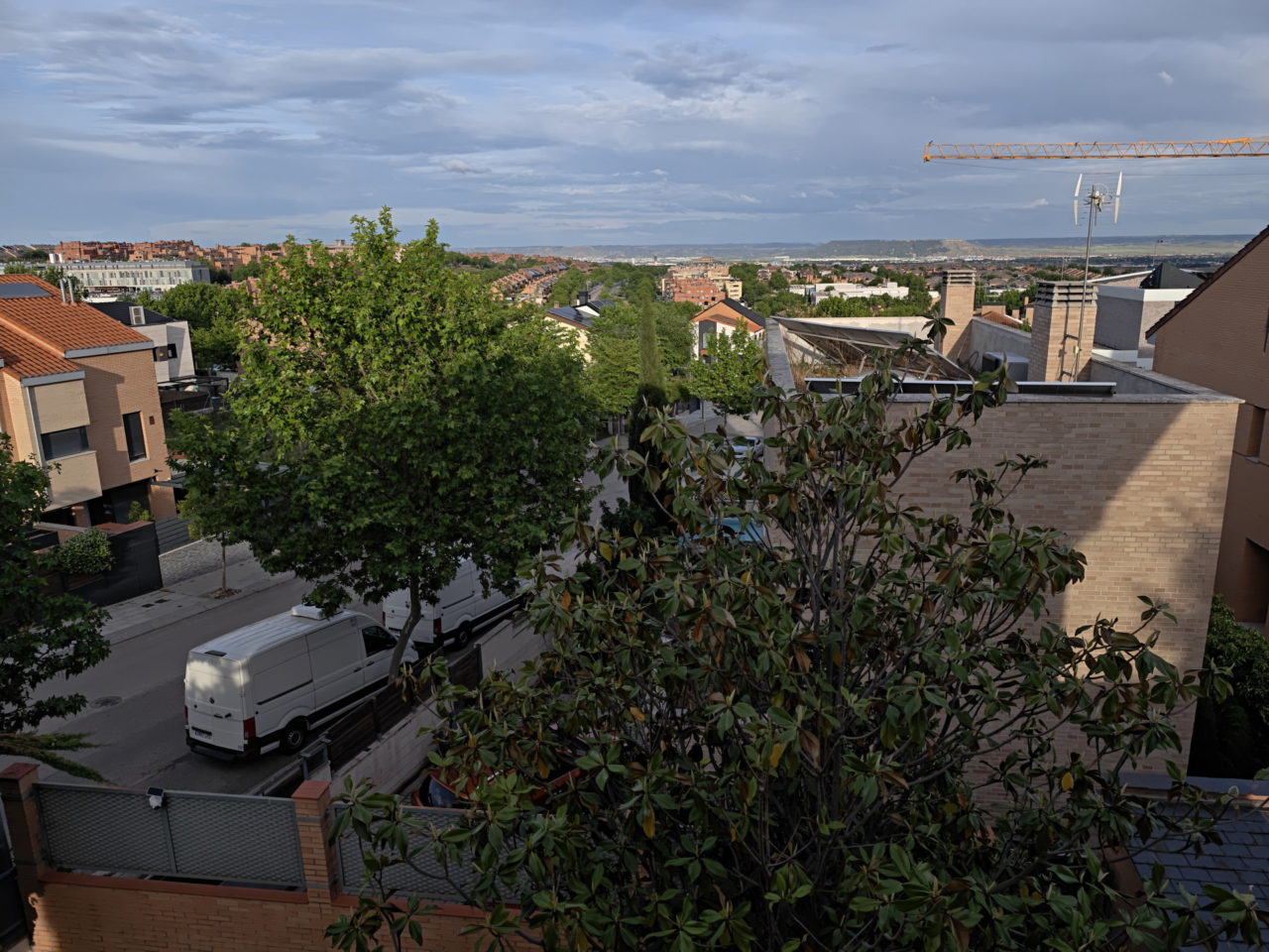 Vista urbana con edificios de ladrillo, árboles verdes y furgonetas estacionadas en una calle soleada.