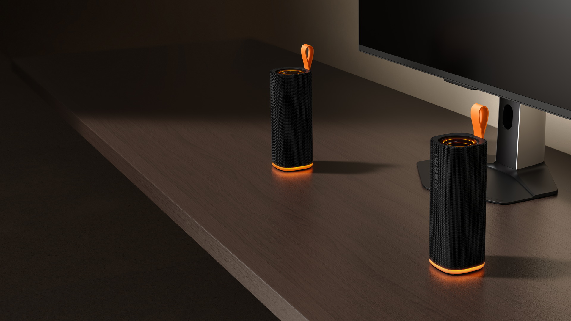 Dos altavoces portátiles negros con detalles naranjas iluminados sobre un escritorio junto a una pantalla.