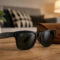 Gafas de sol negras Ray-Ban en mesa de madera, funda marrón y planta decorativa al fondo.
