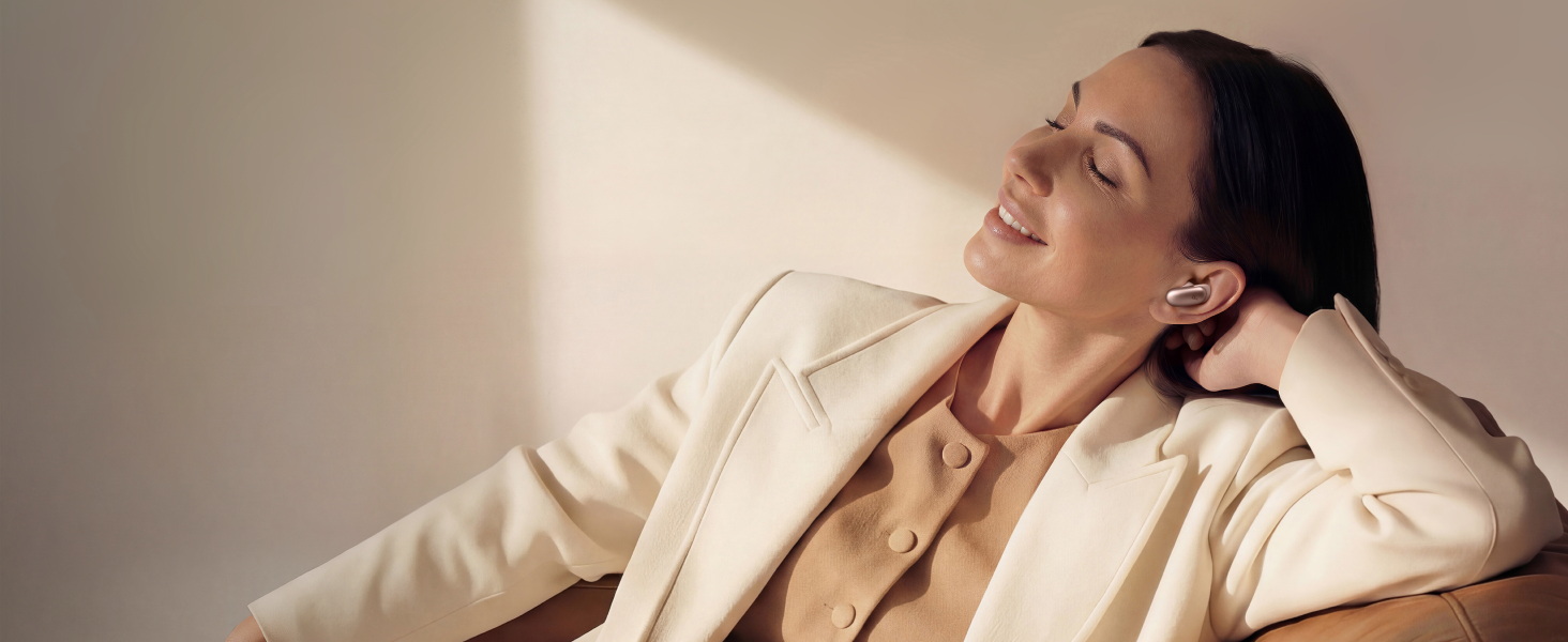 Mujer relajada disfruta de música con auriculares inalámbricos, usando chaqueta beige en ambiente minimalista.
