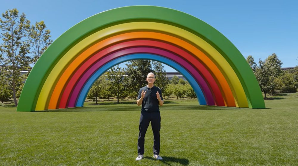 Apple retrasa Siri AI en la Unión Europea para iPhone y iPad por la DMA Escultura de arco iris gigante en un parque verde con cielo despejado y hombre de pie al frente.