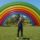 Escultura de arco iris gigante en un parque verde con cielo despejado y hombre de pie al frente.