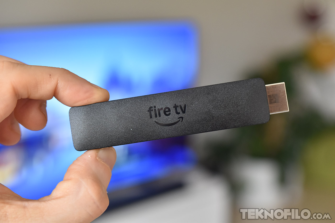 Mano sosteniendo un dispositivo Fire TV Stick frente a una pantalla de televisión borrosa.