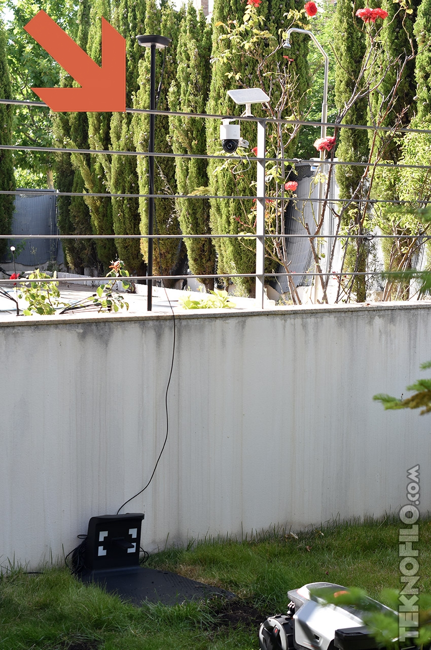 Robots cortacésped en jardín con panel solar y cámaras de seguridad. Flecha señala una instalación en la cámara.