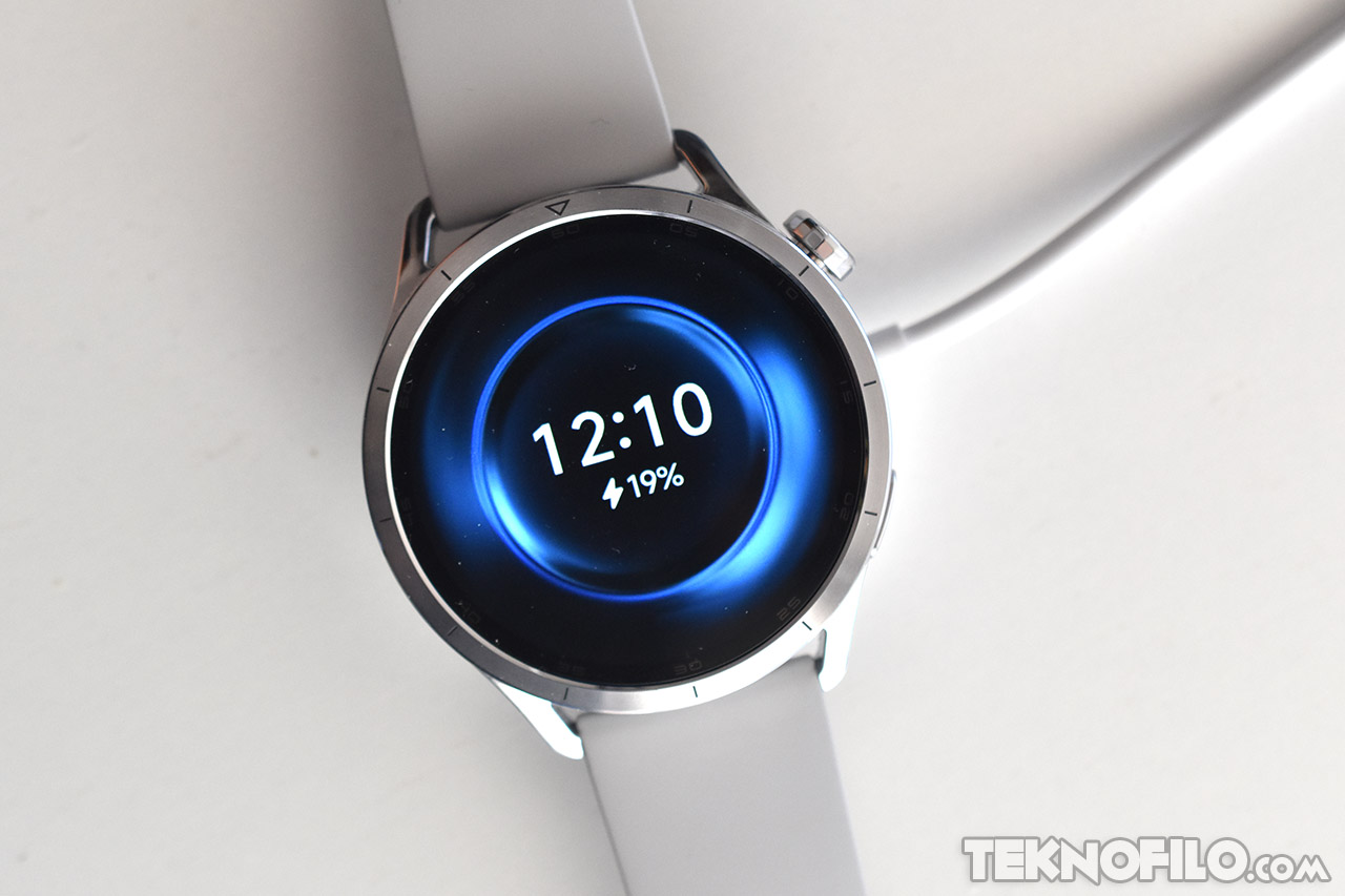 Reloj inteligente plateado con pantalla digital que muestra la hora 12:10 y carga del 19%, sobre fondo blanco.